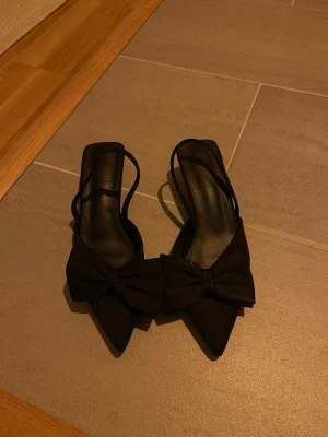 Svarta pumps med rosett och öppen häl - Stilrena svarta pumps med spetsig tå och stor dekorativ rosett framtill. Skorna har öppen häl och tunna remmar som ger en elegant look. Perfekta för att lyfta vilken outfit som helst. Materialet ser ut att vara mocka eller liknande tyg. Aldrig använda 
