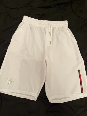 Vita shorts med snörning från Prada - Snygga vita shorts från Prada med elastisk midja och snörning. Shortsen har en diskret logga på vänster ben och en röd detalj på höger sida. Tillverkade i ett lätt och syntetiskt material som passar perfekt till sommaren.