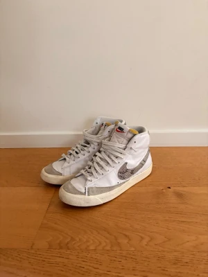 Nike skor - Säljer ett par vita Nike high top sneakers med grått snakeprint på swoosh och häl. Skorna har klassisk retrodesign, vita skosnören och detaljer i mocka på tån och sidan. Perfekta för dig som gillar streetstyle och vill sticka ut lite extra. Hör av er vid minsta fundering! 