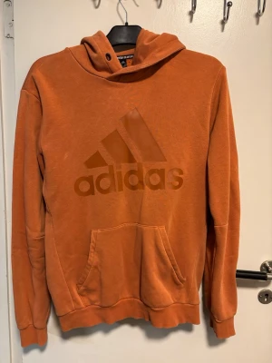 Orange hoodie från Adidas med logga - Säljer en snygg orange hoodie från Adidas med stor logga på bröstet. Hoodien har huva med dragsko, känguruficka och ribbade muddar. Tillverkad i mjukt bomullsmaterial som är skönt att ha på sig. Perfekt för dig som vill ha en sportig och trendig look.