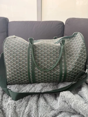 Grön weekend väska  - Snygg grön weekendbag med klassiskt grafiskt mönster och detaljer i mörkgrönt. Väskan har både handtag och justerbar axelrem för smidig användning. Perfekt storlek för kortare resor eller gymmet. Stängs med dragkedja och har silverfärgade metalldetaljer.