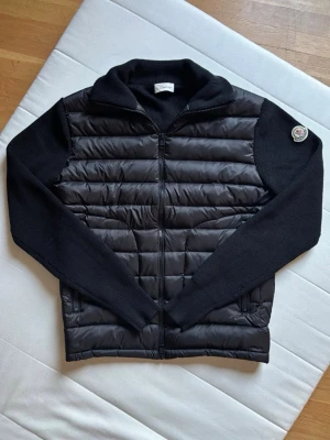 Svart dunväst från Moncler - Snygg svart dunväst från Moncler med stickade ärmar och krage. Västen har quiltad front och rygg i glansigt material, dragkedja framtill och två fickor. Moncler-logga på vänster ärm. Perfekt för lager-på-lager och streetwear.