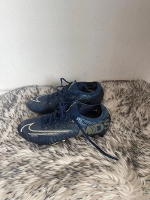 Blå Nike Mercurial fotbollsskor - Säljer ett par blå Nike Mercurial fotbollsskor med vita detaljer och snörning. Skorna har en låg profil och är designade för snabbhet på planen. Snyggt mönster och MDS-tryck på sidan. Perfekta för dig som vill sticka ut på fotbollsplanen.