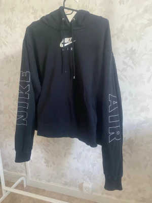 Svart Nike Air hoodie med tryck - Snygg svart hoodie från Nike med stor huva och dragsko. Nike Air-logga på bröstet och stora vita tryck på båda ärmarna där det står 'NIKE' på ena och 'AIR' på andra. Mjuk bomullskänsla och klassisk passform. Perfekt för en sportig och avslappnad stil.
