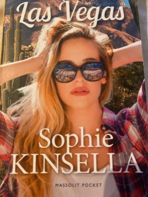 Las Vegas av Sophie Kinsella - Pocketbok med färgstarkt omslag där en ung kvinna med solglasögon och långt blont hår syns mot en ökenbakgrund. Boken är skriven av Sophie Kinsella och utgiven av Massolit Pocket. Perfekt för dig som gillar humoristisk och lättsam skönlitteratur med inslag av äventyr.