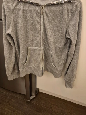 Grå hoodie med dragkedja och paljetter - Grå hoodie med dragkedja framtill och fickor på magen. Huvan är dekorerad med silvriga paljetter för en extra touch. Tillverkad i mjukt sweatshirtmaterial, perfekt för chill dagar. Långa ärmar och ribbade muddar. Storlek 158/164 som xs.