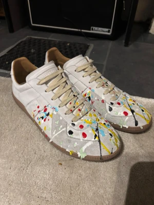 Maison margiela paint splatter gats - Unika vita sneakers från Maison Margiela med färgstänk i rött, blått, gult och svart. Skorna har beige snörning, bruna gummisulor och en klassisk låg siluett. Tillverkade i läder och mocka för en exklusiv känsla och cool look. Storlek 46 men passar som 45 skulle jag säga