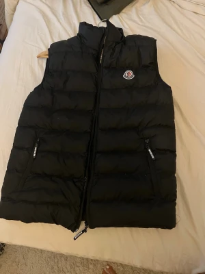 Svart dunväst från Moncler - Snygg svart dunväst från Moncler med hög krage och dragkedja framtill. Västen har två dragkedjeförsedda fickor och Moncler-logga på bröstet. Perfekt för lager-på-lager och håller dig varm med sin fluffiga dunfyllning.