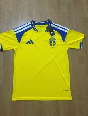 Svensk landslagströja Adidas gul/blå - Säljer en officiell svensk landslagströja från Adidas i gult med blå detaljer och vita ränder på axlarna. Tröjan har broderat Sverige-emblem på bröstet och klassisk Adidas-logga. Tillverkad i lätt och ventilerande funktionsmaterial.
