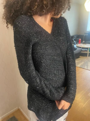 Svart glittrig stickad tröja Milla - Snygg svart stickad tröja från Milla med glittriga inslag. Tröjan har V-ringning, lång ärm och en lite längre passform. Perfekt för dig som vill ha något både bekvämt och trendigt. Materialet är mjukt och stretchigt med en skimrande effekt.perfekt för vintern ifall man vill ha en lång fin kofta