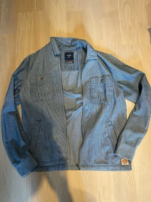 Overshirt från Superdry - Snygg overshirt från Superdry i blått med tunna vita ränder. Jackan har två bröstfickor med knapp, två sidofickor och dragkedja framtill. Tillverkad i ett mjukt, lite tjockare tyg som ger en avslappnad men stilren look.
