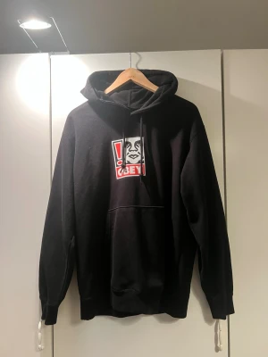 Svart OBEY hoodie med tryck - Svart hoodie från OBEY med stort grafiskt tryck i rött, vitt och svart på bröstet. Original pris 600 kr.