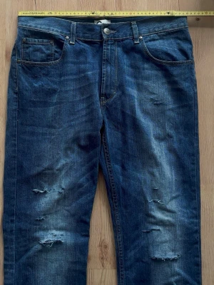 Blå raka jeans - Säljer ett par blå jeans med rak passform och slitna detaljer på både lår och knän. Jeansen har klassisk femficksdesign och är gjorda i denim med snygg tvättad look. Perfekta för en avslappnad och trendig stil.