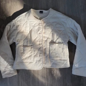 Beige quiltad cardiganjacka - Stilren beige cardiganjacka med quiltat mönster och två stora framfickor. Jackan har rund halsringning, knäppning framtill och är tillverkad i ett mjukt, strukturerat tyg. Perfekt för lager på lager och en avslappnad look.