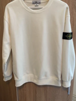 Vit sweatshirt från Stone Island - Vit sweatshirt från Stone Island med klassisk rund halsringning och den ikoniska svarta patchen med logga på vänster ärm. Tröjan har ribbade muddar vid ärmslut och nederkant. Perfekt för en clean och stilren look.