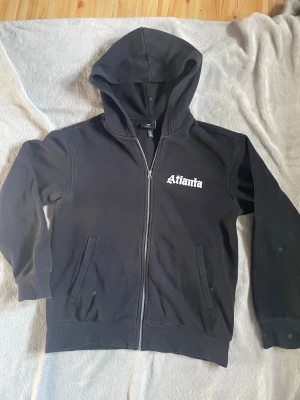Svart hoodie med dragkedja Atlanta - Säljer en svart hoodie från H&M med dragkedja och vit text 'Atlanta' på bröstet. Tröjan har huva, två fickor framtill och ribbade muddar vid ärmar och nederkant. 