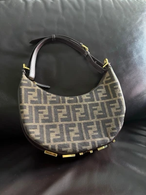  Run Fendi axelremsväska - Säljer en brun handväska från Fendi med klassiskt FF-mönster i beige och brunt. Väskan har en halvmåneform, justerbart axelband i läder och stora guldfärgade bokstäver  FENDI på undersidan. Perfekt för dig som gillar statement-accessoarer.