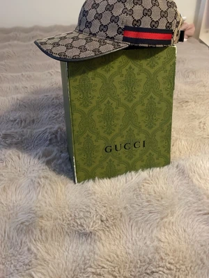 Gucci beige keps med GG-mönster - Säljer en beige keps från Gucci med klassiskt GG-monogram över hela kepsen. Kepsen har en böjd skärm, justerbart spänne bak och en röd-grön rand på sidan. Materialet är canvas med läderdetaljer. Perfekt för dig som vill ha en lyxig och trendig accessoar.