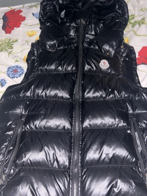 Svart dunväst från Moncler - Snygg svart dunväst från Moncler med glansig finish och klassisk logga på bröstet. Västen har dragkedja framtill, två dragkedjefickor och hög krage. Perfekt för lager-på-lager och håller dig varm med dunfyllning.