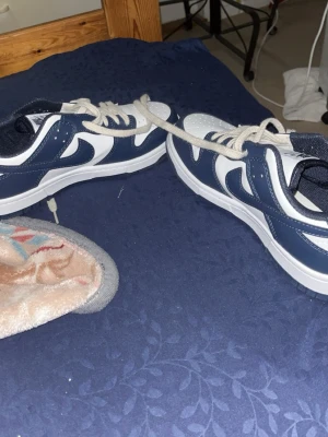 Nike Dunk Low blå och vita sneakers - Snygga Nike Dunk Low sneakers i blått och vitt med klassisk swoosh på sidan. Skorna har vita snören, perforerad tåbox och en robust gummisula. Perfekt för dig som vill ha en stilren och sportig look till vardagsoutfiten.