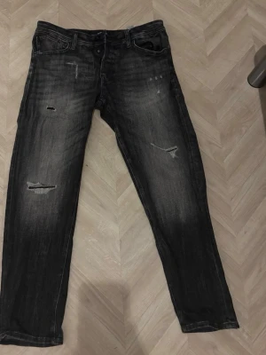 Svarta slitna jeans med raka ben - Snygga svarta jeans med slitningar och hål på benen för en trendig look. Jeansen har raka ben, klassisk femficksmodell och tydliga tvättade detaljer. Perfekta för dig som gillar en avslappnad och edgy stil. 32 i midjan och 30 i längden 