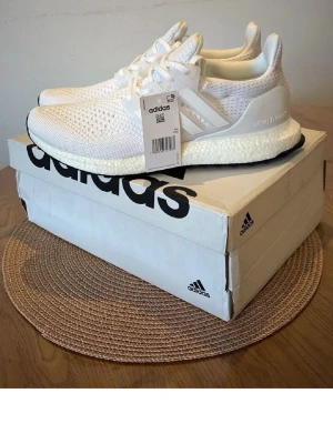 Adidas Ultra Boost vita sneakers - Fräscha vita Adidas Ultra Boost sneakers med stickad ovandel och klassiska tre ränder på sidan. Skorna har en bekväm Boost-sula och ventilerande meshmaterial. Perfekta för dig som gillar stilrena och sportiga sneakers.