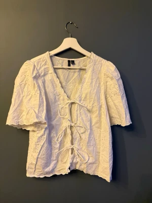 Vit blus med knytning och broderi - Söt vit blus med broderade blommor och hålmönster. Blusen har v-ringning, knytning framtill med tre rosetter och korta ärmar med lätt puff. Tillverkad i ett tunt, luftigt tyg. 💕