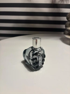 Diesel Only The Brave parfym - Aldrig använd. Cool parfymflaska från Diesel formad som en knytnäve i mörkgrått glas med silverfärgat lock. Flaskan har en unik och edgy design med Diesel-loggan i metall på sidan. Perfekt för dig som gillar statement-detaljer på parfymhyllan.