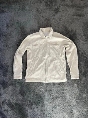 Vit overshirt i manchester - Säljer en stilren vit overshirt från Vailent i mjuk manchester. Jackan har två stora bröstfickor, dragkedja framtill och klassisk krage. Perfekt lager på lager-plagg för dig som gillar minimalistisk stil.