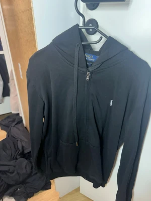 Svart hoodie från Polo Ralph Lauren - Snygg svart hoodie från Polo Ralph Lauren med dragkedja framtill, huva med dragsko och klassisk logga broderad på bröstet. Tillverkad i mjukt material och har en stor ficka framtill. Perfekt för en chill och stilren look.