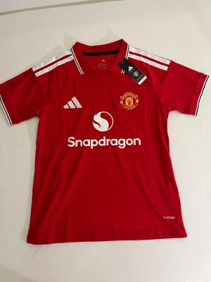 Manchester United matchtröja röd - Snygg röd Manchester United matchtröja från Adidas med vita detaljer och klubbmärke på bröstet. Tröjan har korta ärmar, vit logga och sponsortryck på framsidan samt klassiska Adidas-ränder på axlarna. Perfekt för fotbollsträning eller att bära till vardags.