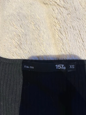 Svart ribbad långärmad topp 157 - En svart, ribbad långärmad topp från 157 i storlek XS. Toppen har en enkel och stilren design med rund halsringning och smal passform. Perfekt att styla med jeans eller kjol för en clean look.