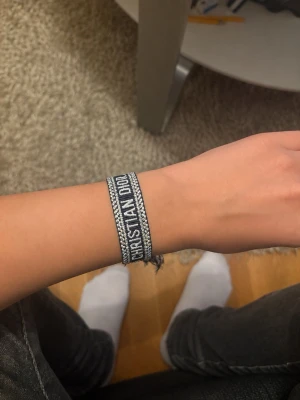 Broderat armband Christian Dior - Broderat armband från Christian Dior i svart och vitt med tydlig logga. Armbandet har justerbar knytning med tofsar och ett mönstrat band. Perfekt accessoar för att lyfta din stil med en lyxig touch.