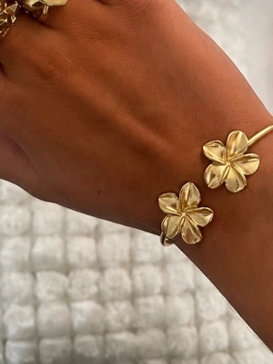 Guldigt armband med blommor - Snyggt armband i guldfärgad metall med två stora blommor som dekoration. Blommorna har fem blad och ger en trendig och elegant look. Perfekt för dig som vill ha en unik accessoar som sticker ut.
