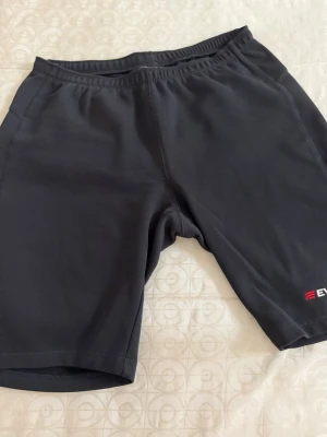 Svarta träningsshorts i syntet - Säljer ett par svarta träningsshorts i syntetmaterial med elastisk midja. De har en enkel och sportig design med en liten logga i rött och vitt nere vid benet. Perfekta för gymmet eller löprundan!