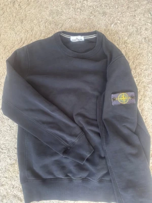 Svart sweatshirt från Stone Island - Svart sweatshirt från Stone Island med klassisk rund hals och ikonisk kompasspatch på vänster ärm. Tröjan har ribbade muddar vid ärmslut och nederkant. Perfekt för en clean och stilren look. Fin äkta stone Island tröja, storlek L men sitter som M
