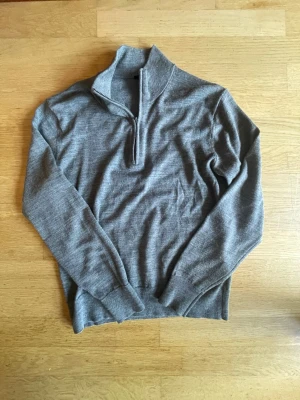 Grå stickad halfzip  - En stilren grå stickad tröja med halv dragkedja i halsen. Tröjan har långa ärmar, ribbstickade muddar och en hög krage som kan vikas ner. Perfekt för lager-på-lager och enkel att matcha med jeans eller kjol. Materialet är merinoull