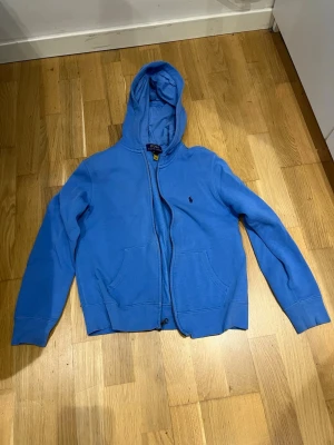 Blå zip hoodie från Polo Ralph Lauren - Blå hoodie med dragkedja från Polo Ralph Lauren. Klassisk modell med huva, två fickor framtill och det ikoniska lilla broderade logot på bröstet. Perfekt för en avslappnad och stilren look. Mjuk bomullskänsla och ribbade muddar vid ärmar och midja.