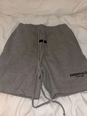 Grå Essentials shorts med snörning - Säljer ett par grå shorts från Fear of God Essentials med långa snören i midjan och tryckt logga på benet. De har en relaxed fit och är perfekta för chill dagar. Tillverkade i mjuk bomull för maximal komfort.