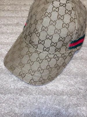 Beige keps med GG-mönster från Gucci - Snygg beige keps från Gucci med det klassiska GG-monogrammet över hela kepsen. På sidan finns en grön och röd rand som ger extra lyxig känsla. Kepsen har böjd skärm och är tillverkad i ett slitstarkt tygmaterial.