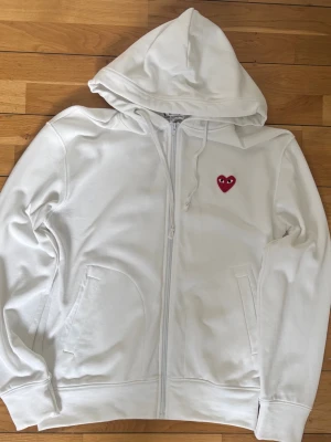 Vit hoodie med rött hjärta - Säljer en vit hoodie med dragkedja och huva. På bröstet sitter ett rött hjärta med ögon, vilket ger en unik och lekfull touch. Hoodien har fickor på sidorna och ribbade muddar vid ärmslut och nederkant. Perfekt för dig som gillar stilrena plagg med en twist.