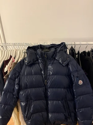 Mörkblå dunjacka från Moncler - Snygg mörkblå dunjacka från Moncler med glansig finish och avtagbar huva. Jackan har två dragkedjeförsedda fickor fram och Moncler-logga på ärmen. Insidan är beige och jackan stängs med dragkedja. Perfekt för kalla vinterdagar. Inga defekter och pris kan diskuteras vid snabb affär 