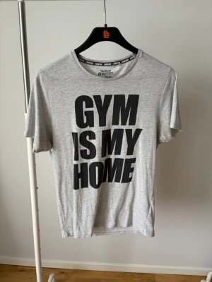 Grå t-shirt med tryck 'GYM IS MY HOME' - Snygg grå t-shirt med stort svart tryck 'GYM IS MY HOME' på framsidan. Klassisk rund hals och korta ärmar. Perfekt för dig som älskar gymmet och vill ha en bekväm och stilren look. Tillverkad i mjuk bomull.