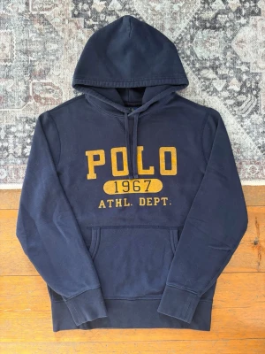 Graphic Ralph Lauren Hoodie - Storlek S - Tjena! 👋 Säljer en riktigt fet mörkblå Polo Ralph Lauren hoodie i storlek S med det klassiska trycket ”POLO 1967 ATHL. DEPT.”. 🎽✨ Tröjan är i mycket gott skick. Trycket har blivit lite naturligt luddigt (se bilder), vilket bara ger en skönare vintage-look! 100% äkta har ni några fårgor är det bara att höra av sig 😘