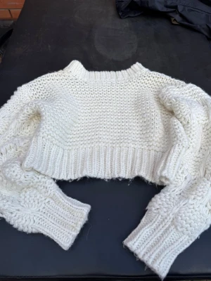 Vit cropped stickad tröja Miss Lisibell - Säljer en chunky vit cropped stickad tröja från Miss Isabel. Tröjan har grovstickat mönster, bred ribbad kant och långa ärmar med flätstickade detaljer. Perfekt för lager-på-lager och en cool look till jeans eller kjol.