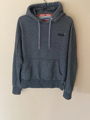 Mörkgrå hoodie från Superdry - Mörkgrå hoodie från Superdry med broderad logga på bröstet. Klassisk känguruficka framtill, ribbade muddar och justerbar huva med dragsko. Mjuk och skön bomullsblandning, perfekt för chill dagar.