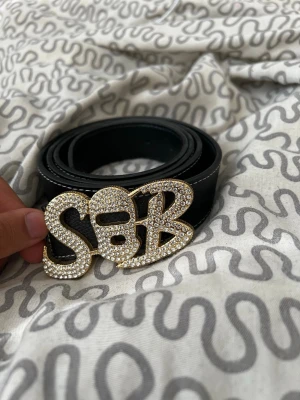 Svart bälte med glittrigt S&B-spänne - Snyggt svart bälte med ett stort, glittrigt S&B-spänne i guld och strass. Bältet har en klassisk form och är gjort i ett material som liknar skinn. Perfekt för att ge outfiten lite extra bling.