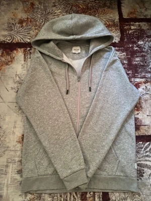 Grå zip hoodie från Jack & Jones - En stilren grå hoodie från Jack & Jones med dragkedja framtill och två stora fickor. Klassisk huva och mjukt material som känns skönt mot huden. Perfekt för en avslappnad look och enkel att matcha med jeans eller joggers.
