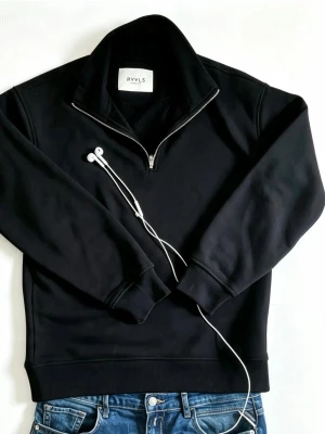 Svart half-zip hoodie från RYVLS - Svart hoodie från RYVLS med halv dragkedja och hög krage. Tillverkad i mjukt material med ribbade muddar vid ärmslut och nederkant. Enkel och stilren design som passar perfekt till jeans eller joggers.