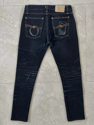 Nudie Jeans 31/32 - Säljer ett par mörkblå jeans från Nudie Jeans med skön passform och snygga slitningar på framsidan. Jeansen har klassiska fem fickor, kontrastsömmar och Nudie-logga på bakfickan. Perfekta för en trendig och avslappnad stil.